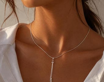 May include: A delicate silver-toned necklace featuring a thin, smooth chain and a vertical drop pendant. The pendant consists of two slender silver strands, each accented with a small, polished silver bead at the bottom, creating a refined look.