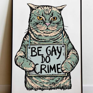 May include: A framed art print featuring a cartoon cat with a stern expression. The cat, in shades of green and tan, holds a sign that reads "BE GAY DO CRIME" in black text. The artwork has a vintage, hand-drawn style.