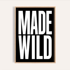 以下が含まれることがあります： 「MADE WILD」の文字が白抜きの太字で黒い背景にプリントされた額装アート。 フレームは自然な木の色で、全体的にモダンでミニマリストなデザインです。