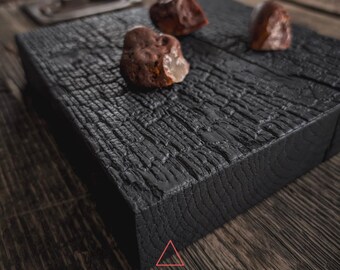 May include: A black wooden serving board with a rough, textured surface. Four small, reddish-brown stones are arranged on the board.