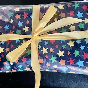 May include: A gift wrapped in clear plastic with a blue background fabric with colourful stars. The gift is tied with a yellow ribbon.