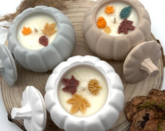 May include: Three ceramic pumpkin-shaped candle holders with lids. Each holder has a white candle with fall-themed decorations, including leaves and acorns.