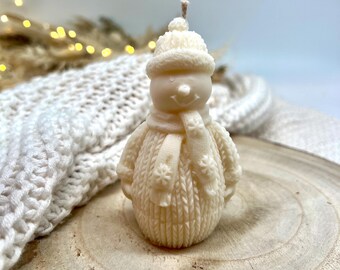 May include: A white snowman candle with a knitted hat and scarf. The candle is sitting on a wooden surface.