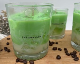 May include: Three clear glass candles with a layered design, resembling an iced matcha latte. The top layer is bright green, the bottom is white, and the text "Iced Matcha Latte" is visible on the front of the glass. Coffee beans are scattered around the candles.