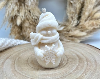 May include: A white snowman candle with a scarf and a snowflake in its hands. The candle is sitting on a wooden surface.