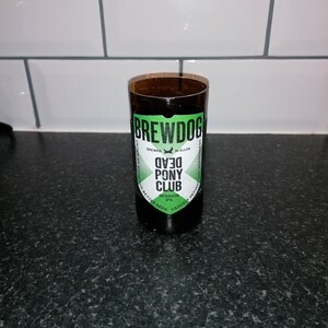 Puede incluir: Una botella de vidrio marrón con una etiqueta verde que dice "Brewdog Dead Pony Club Session IPA". La etiqueta también incluye el texto "Brewed in Ellon, Scotland" y "Fiercely Defiant and Independent".