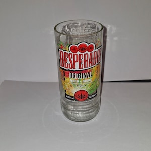 Könnte beinhalten: Ein klares Bierglas mit dem "DESPERADOS"-Logo in Rot und Weiß. Das Glas hat eine zylindrische Form und einen leicht ausgestellten Boden. Das Glas ist 13 cm hoch und fasst 330 ml.