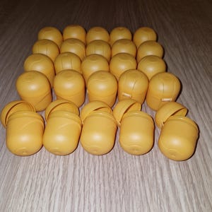 Puede incluir: Una colección de juguetes de cápsulas de plástico amarillas, algunas abiertas y otras cerradas, dispuestas sobre una superficie de grano de madera clara. Las cápsulas tienen forma ovalada con una tapa con bisagras y son de un color uniforme.