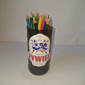 Puede incluir: Un recipiente de vidrio marrón hecho a partir de una botella de cerveza Żywiec, con una etiqueta blanca que muestra un logotipo rojo y la imagen de dos figuras con vestimenta tradicional. El recipiente contiene lápices de madera de colores.