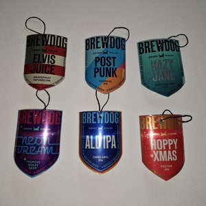 Op de afbeelding: Zes verschillende Brewdog-bierfleshangers, elk met een andere biernaam en ontwerp. De hangers zijn schildvormig en hebben een zwart touwtje om op te hangen. De bieren zijn: Elvis Juice, Post Punk, Hazy Jane, Neon Dream, Ald IPA en Hoppy Xmas.