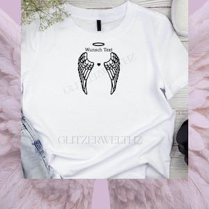 Könnte beinhalten: Weißes T-Shirt mit einem schwarzen Engelsflügel-Design, einem Heiligenschein und dem Text "Wunsch Text". Ein kleines schwarzes Herz befindet sich zwischen den Flügeln. Das Shirt hat einen Rundhalsausschnitt und kurze Ärmel.