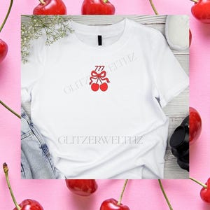 Könnte beinhalten: Weißes T-Shirt mit rotem Kirschdesign und der Zahl 777. Das Shirt befindet sich auf einem rosa Hintergrund mit frischen Kirschen an den Rändern. Der Text "GLITZERWELTHZ" ist auf dem Shirt aufgedruckt.