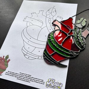 May include: A stained glass heart-shaped ornament with red, green, and clear glass, and a black border. A printed pattern of a heart with numbered sections is in the background. The ornament is hanging by a black cord.