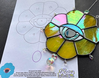May include: A stained glass flower-shaped suncatcher with a central eye design, featuring yellow, green, pink, and blue iridescent glass. A pattern for the suncatcher is visible in the background. The suncatcher has a decorative crystal pendant.