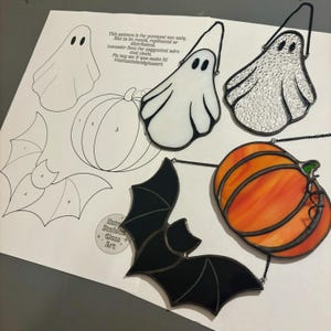 May include: A set of stained glass Halloween decorations, including a ghost, a bat, a pumpkin, and a ghost. The decorations are all outlined in black and have a variety of colours. The decorations are hanging from chains.