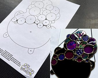 Puede incluir: Una pieza de vidriera en forma de caldero negro con una parte superior burbujeante y colorida, junto a un patrón de papel. Las burbujas de vidrio presentan tonos de morado, azul y verde. El patrón de papel tiene círculos numerados.