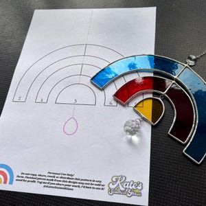 May include: A stained glass rainbow suncatcher with blue, red, and yellow segments, hanging from a silver chain. A pattern for the suncatcher is on a white paper with the text "Kate's Stained Glass".