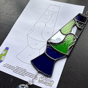 Pode incluir: Um design de lâmpada de lava em vitral em tons de azul, verde e branco, com uma moldura de metal prateado. O design está ao lado de um padrão de papel com seções numeradas. O texto "Kate's Stained Glass" é visível.