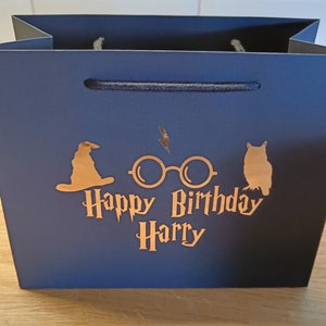 May include: A dark blue gift bag with a gold foil design featuring Harry Potter themed icons, including a wizard's hat, glasses, and an owl. The text "Happy Birthday Harry" is also included in gold foil.