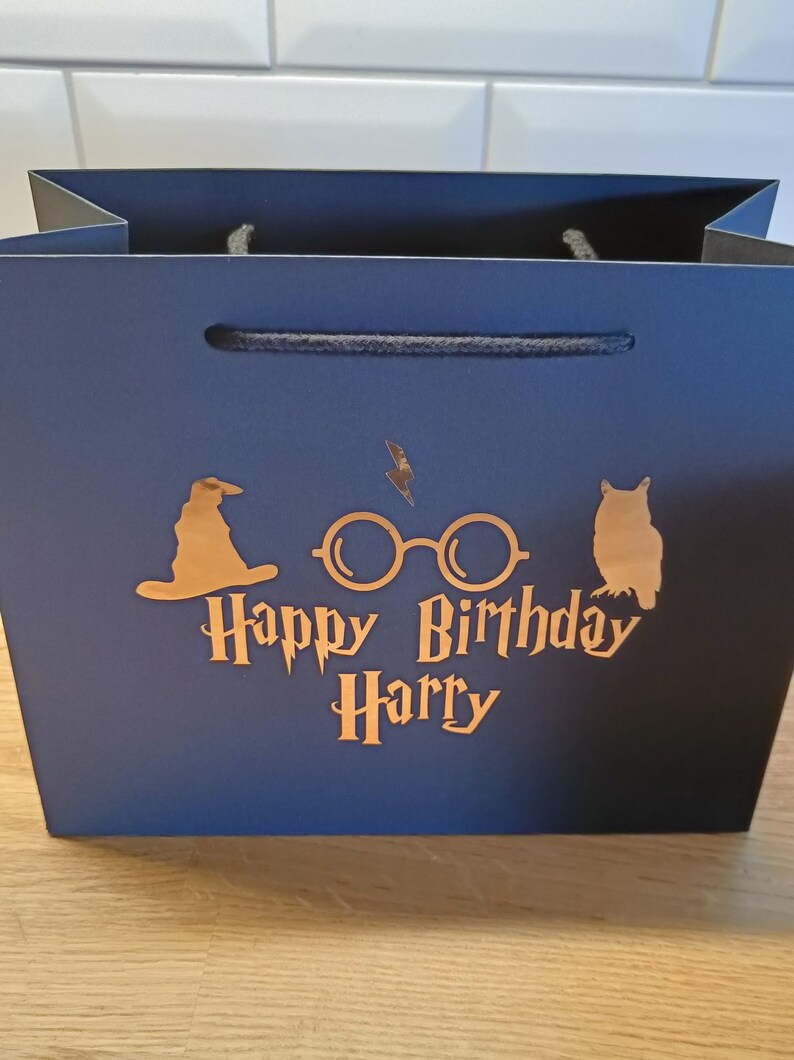 May include: A dark blue gift bag with a gold foil design featuring Harry Potter themed icons, including a wizard's hat, glasses, and an owl. The text "Happy Birthday Harry" is also included in gold foil.