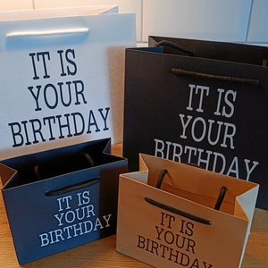 May include: Four gift bags with the text "It is your birthday" printed on them. The bags are in different colors: white, black, black, and brown.