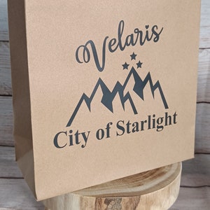 May include: A brown paper gift bag with a red rope handle. The bag has a black graphic design with the text "Velaris City of Starlight".