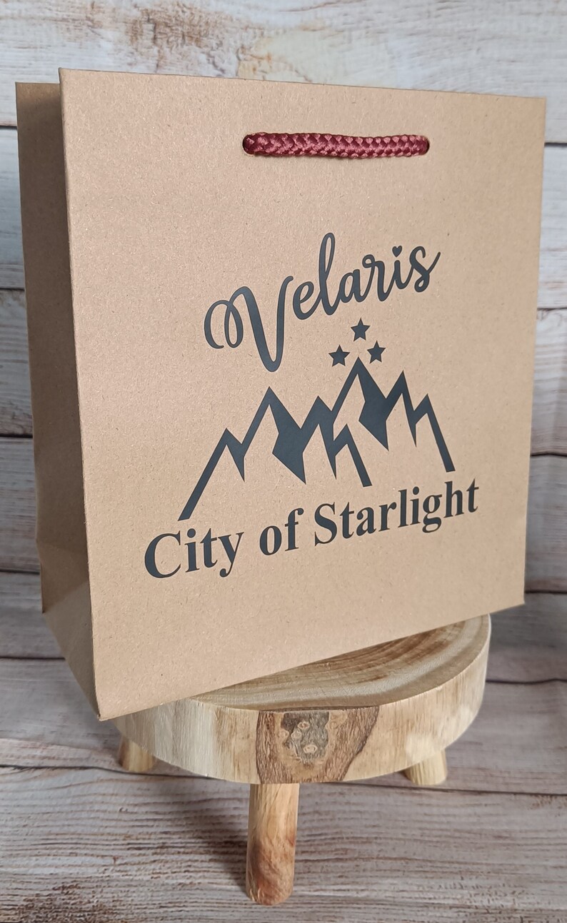May include: A brown paper gift bag with a red rope handle. The bag has a black graphic design with the text "Velaris City of Starlight".
