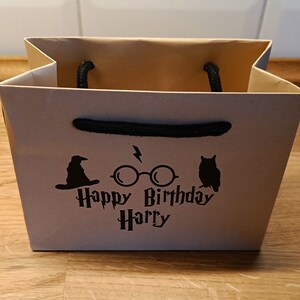 May include: A brown paper gift bag with black handles. The bag has a black graphic of a wizard's hat, glasses with lightning bolts, and an owl. The text "Happy Birthday Harry" is printed in black.