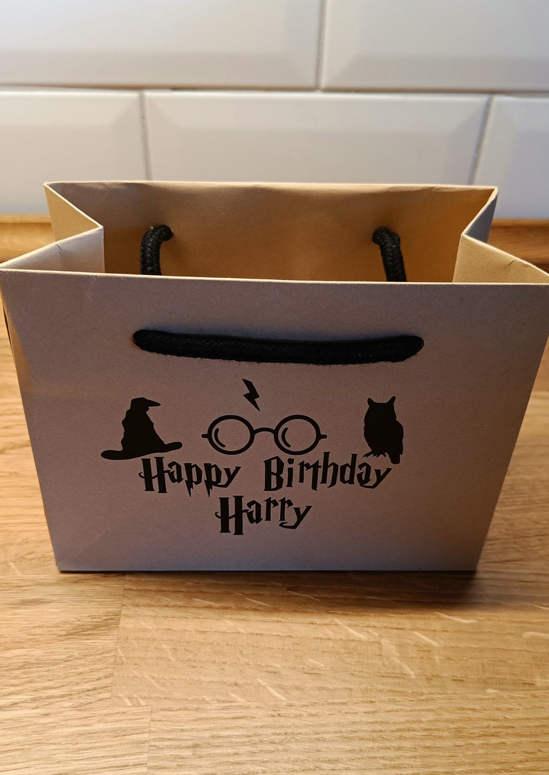 May include: A brown paper gift bag with black handles. The bag has a black graphic of a wizard's hat, glasses with lightning bolts, and an owl. The text "Happy Birthday Harry" is printed in black.