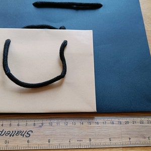 May include: Two paper shopping bags, one beige and one dark blue, with black rope handles. The beige bag has a black rope handle that forms a U-shape. The bags are sitting on a wooden surface.