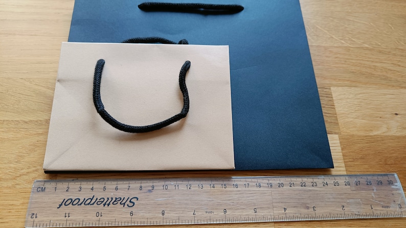 May include: Two paper shopping bags, one beige and one dark blue, with black rope handles. The beige bag has a black rope handle that forms a U-shape. The bags are sitting on a wooden surface.
