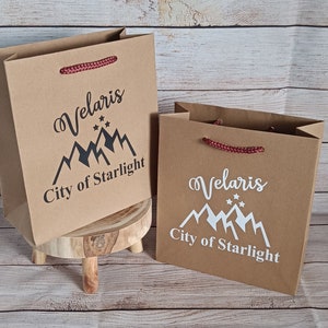 May include: Two brown paper gift bags with red handles. Both bags have a white mountain range design with stars and the text "Velaris City of Starlight".