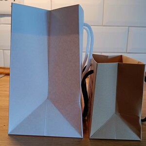 May include: Two paper shopping bags, one white and one brown, are side by side on a wooden surface. The white bag has white handles and the brown bag has black handles.