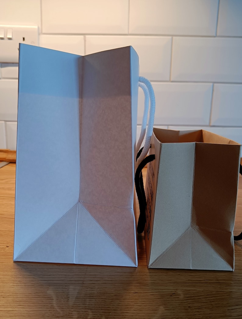 May include: Two paper shopping bags, one white and one brown, are side by side on a wooden surface. The white bag has white handles and the brown bag has black handles.