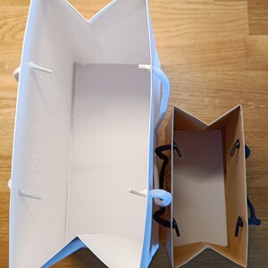 May include: Two white paper gift bags with black handles. The larger bag is open and the smaller bag is closed.
