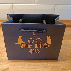 May include: Black paper gift bag with a gold design featuring a wizard's hat, glasses, and an owl. The text "Happy Birthday Harry" is printed on the bag.