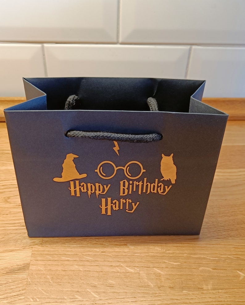 May include: Black paper gift bag with a gold design featuring a wizard's hat, glasses, and an owl. The text "Happy Birthday Harry" is printed on the bag.