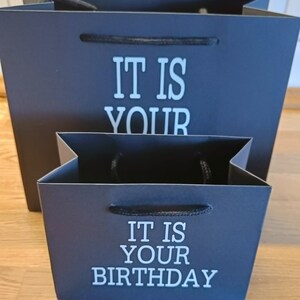 May include: Two black gift bags with white text. The larger bag says "It is your" and the smaller bag says "It is your birthday".