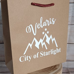 May include: A brown paper gift bag with a red rope handle. The bag has a white design of mountains and stars with the text "Velaris City of Starlight".