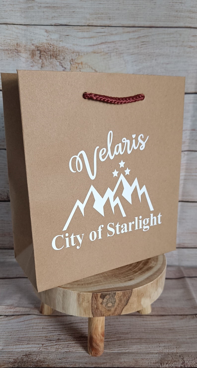 May include: A brown paper gift bag with a red rope handle. The bag has a white design of mountains and stars with the text "Velaris City of Starlight".