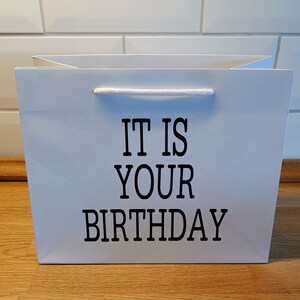 May include: White gift bag with black text that reads "It is your birthday".