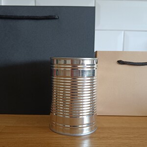 May include: Two paper gift bags, one black and one brown, with black handles. A silver can sits in front of the bags.
