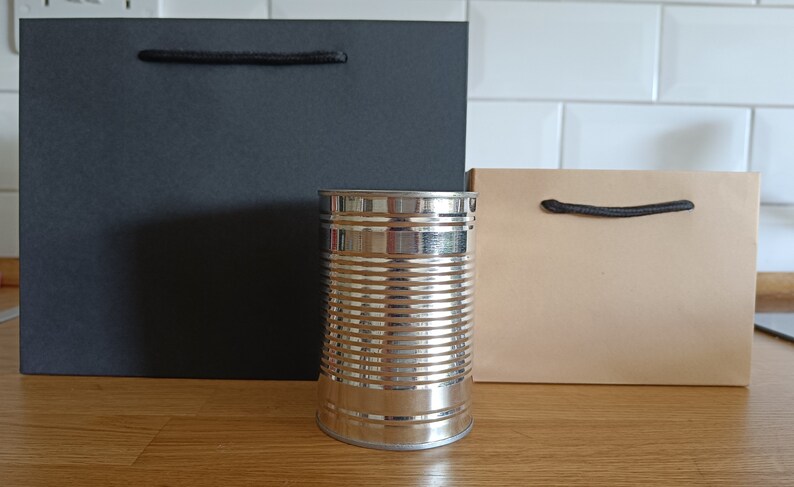 May include: Two paper gift bags, one black and one brown, with black handles. A silver can sits in front of the bags.