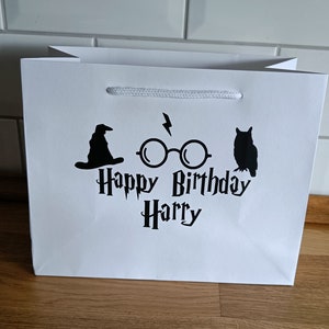 May include: White gift bag with a black graphic of a wizard's hat, glasses, and an owl. The text "Happy Birthday Harry" is printed on the bag.