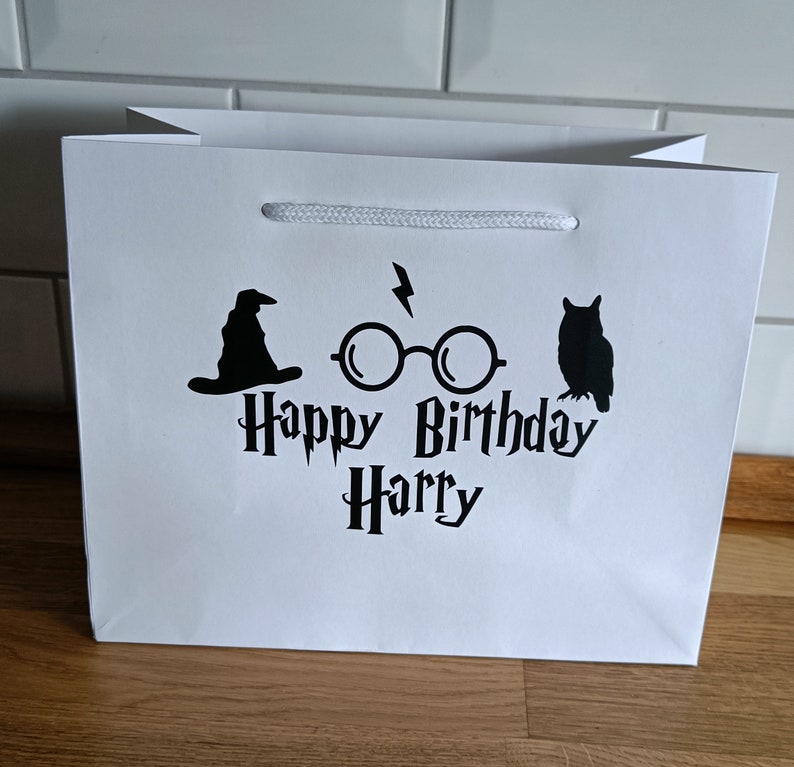 May include: White gift bag with a black graphic of a wizard's hat, glasses, and an owl. The text "Happy Birthday Harry" is printed on the bag.