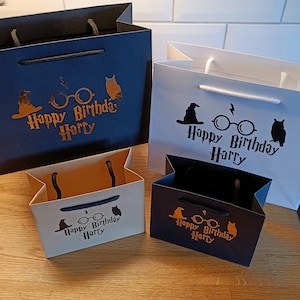 May include: Four gift bags in different sizes and colors. The bags are decorated with a Harry Potter theme, including a black silhouette of a witch's hat, glasses, and an owl. The text on the bags reads "Happy Birthday Harry" in gold lettering.