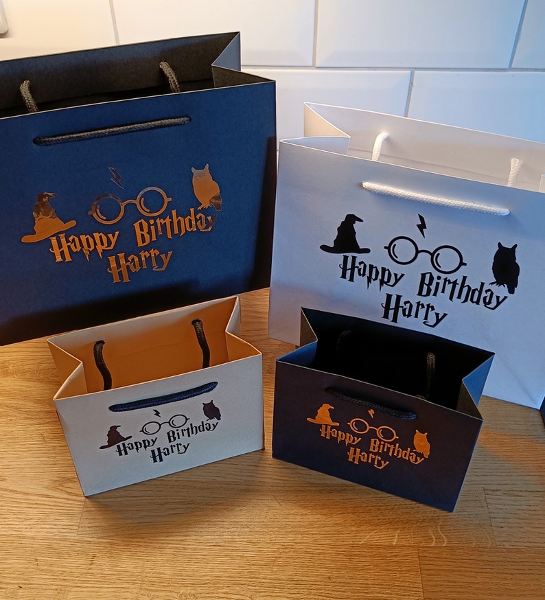 May include: Four gift bags in different sizes and colors. The bags are decorated with a Harry Potter theme, including a black silhouette of a witch's hat, glasses, and an owl. The text on the bags reads "Happy Birthday Harry" in gold lettering.