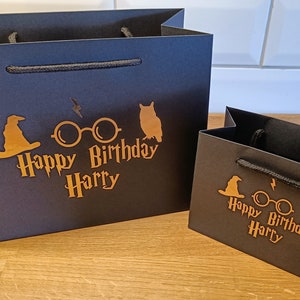May include: Two black gift bags with gold lettering and Harry Potter themed graphics. The larger bag has a wizard's hat, glasses, and an owl. The smaller bag has a wizard's hat, glasses, and an owl. Both bags say "Happy Birthday Harry".