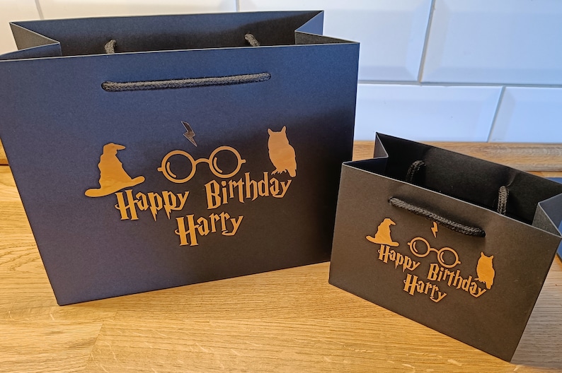 May include: Two black gift bags with gold lettering and Harry Potter themed graphics. The larger bag has a wizard's hat, glasses, and an owl. The smaller bag has a wizard's hat, glasses, and an owl. Both bags say "Happy Birthday Harry".