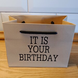 May include: A brown paper gift bag with black handles and the text "IT IS YOUR BIRTHDAY" printed on the front.